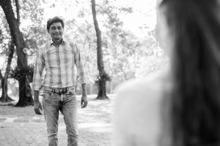 Happy Indian man smiling and standing while looking at beautiful woman in love at peaceful green parkの写真素材