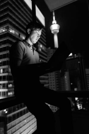 Young handsome man using laptop against glass window with view of the city at nightの写真素材
