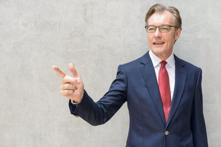 Happy mature businessman in suit pointing finger and directing against concrete wall