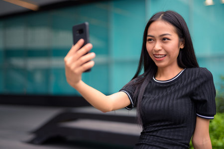 Happy young beautiful Asian tourist woman taking selfie at the mall outdoors
