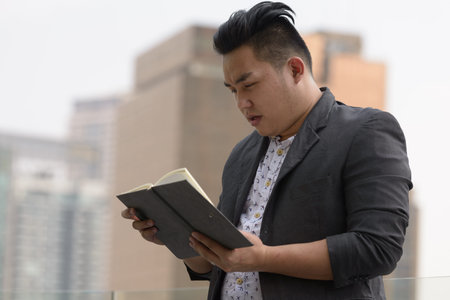 Young overweight Asian businessman reading book in the city