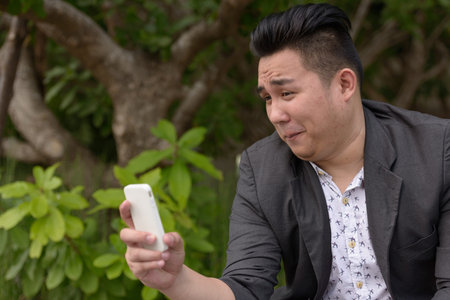 Young overweight Asian businessman using phone at the park