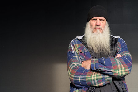 Mature bearded hipster man looking serious with arms crossed against black wall