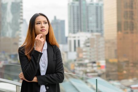 Young beautiful Asian businesswoman thinking against view of the cityの写真素材