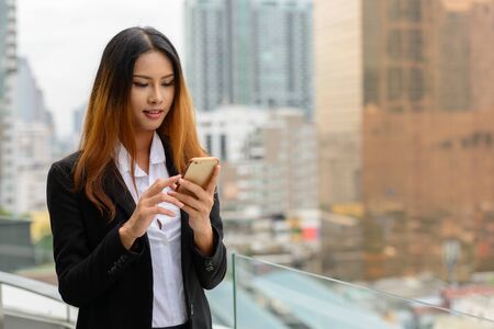 Happy young beautiful Asian businesswoman using phone against view of the cityの写真素材