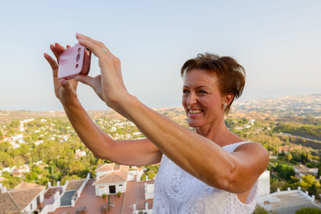 Happy mature tourist woman taking picture with phone of the beautiful scenery of Mijas village in...