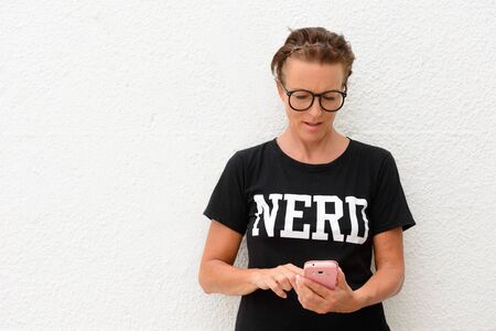 Mature nerd woman wearing big eyeglasses and standing against white background outdoors while using mobile phone and touching screenの写真素材