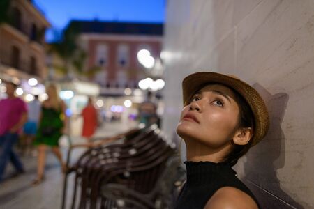 Young beautiful Asian tourist woman thinking and sitting outdoors at nightの写真素材