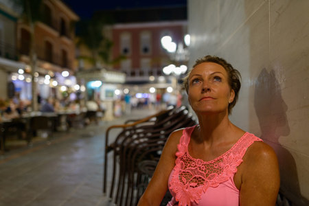Mature beautiful tourist woman sitting and thinking outdoors at nightの写真素材