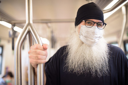 Face of mature bearded hipster man thinking with mask for protection from corona virus outbreak inside the trainの写真素材