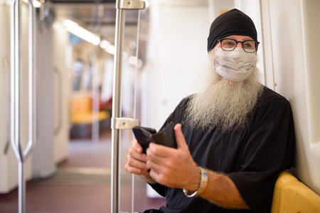 Mature bearded hipster man with mask thinking while using phone inside the train