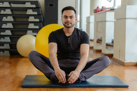Full body shot of young bearded Indian man stretching legs at the gym