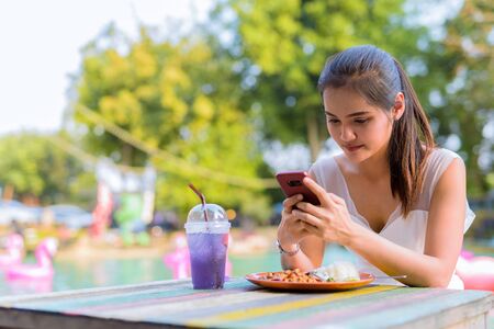 Young beautiful Asian woman using phone while sitting outdoorsの写真素材