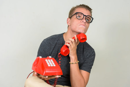 Young blond nerd man with eyeglasses thinking while using old telephone