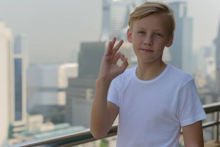 Young blond handsome boy giving ok sign against view of the cityの写真素材