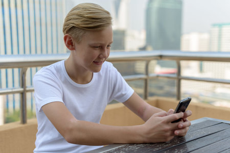 Happy young handsome boy with blond hair sitting while using phone outdoors
