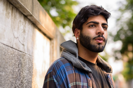 Face of young handsome bearded Indian hipster man thinking in the streets