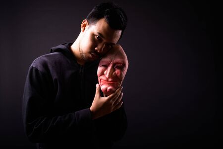Portrait of crazy young Asian man with hoodie holding horror maskの写真素材