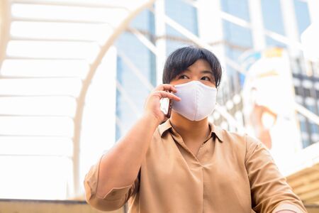 Overweight Asian woman with mask talking on the phone while sitting in the cityの写真素材