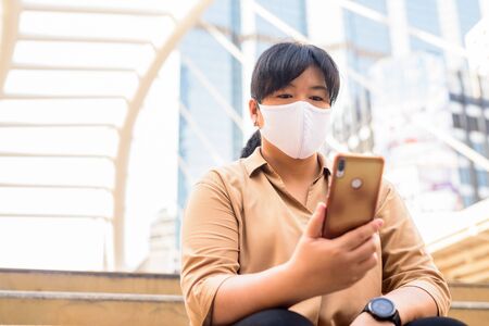 Overweight Asian woman with mask using phone while sitting in the cityの写真素材