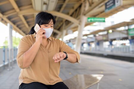 Overweight Asian woman with mask talking on the phone and checking time at the sky train stationの写真素材