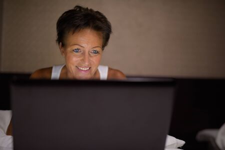 Happy mature beautiful tourist woman using laptop in the bedroomの写真素材