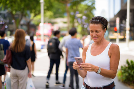 Happy mature beautiful tourist woman using phone in the city streets outdoorsの写真素材