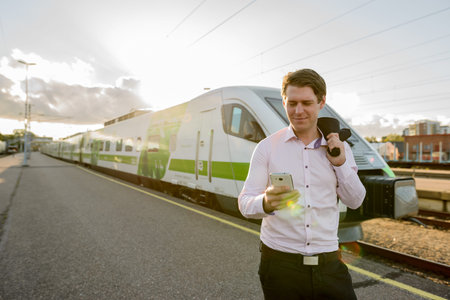 Young happy businessman smiling and using mobile phone in front of train at railway station
