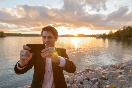 Young happy businessman smiling while taking pictures with mobile phone in beautiful scenic shore