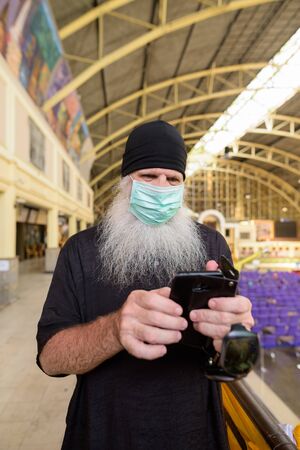 Mature bearded tourist man using phone with mask for protection from corona virus outbreak at railway stationの写真素材