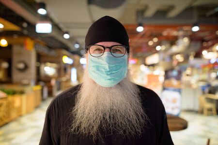 Mature bearded hipster man with mask for protection from corona virus outbreak at the mallの写真素材