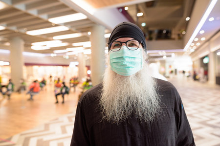 Mature bearded hipster man with mask for protection from corona virus outbreak shopping with...