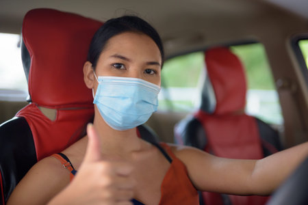 Young Asian woman with mask for protection from corona virus outbreak giving thumbs up inside the...