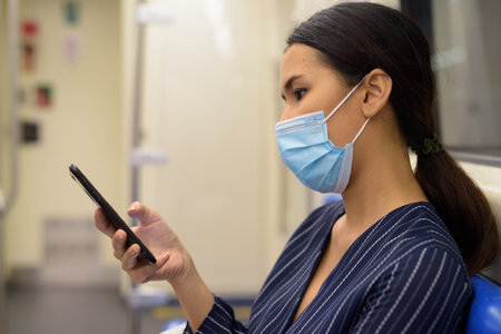 Face of young Asian businesswoman with mask using phone and sitting with distance at the subway...