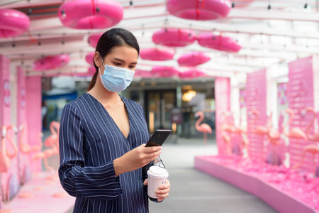 Young Asian businesswoman with mask using phone while having coffee on the go as new normal in the...