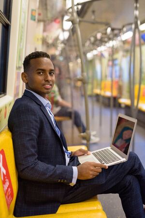Full body shot of happy young handsome African businessman using laptop while sitting with distance inside the trainの写真素材