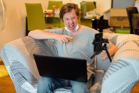 Happy young man with long hair using laptop and vlogging in quarantine at homeの写真素材
