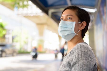 Face of young Asian woman with mask thinking while sitting at the bus stopの写真素材