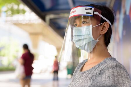 Face of young Asian woman wearing mask and face shield while sitting at the bus stopの写真素材