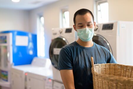 Young Asian man with mask for protection from corona virus outbreak at the laundromat washing clothesの写真素材