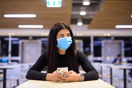 Young Indian woman with mask thinking while using phone and sitting with distance at food courtの写真素材
