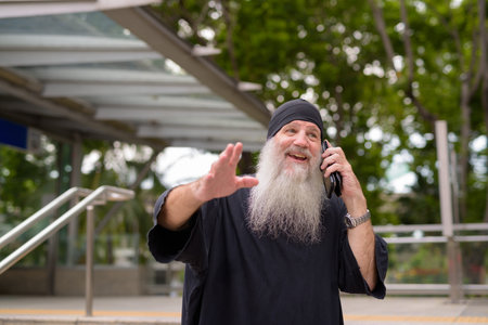 Happy mature handsome bearded hipster man talking on the phone and waving hand outside the subway train stationの写真素材
