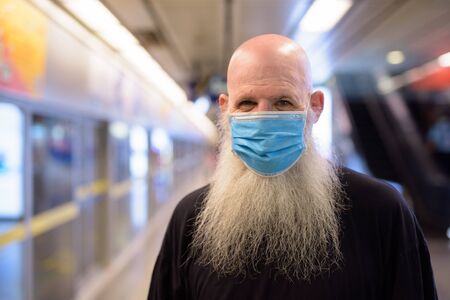 Face of mature bald bearded man wearing mask for protection from corona virus outbreak at the subway train stationの写真素材