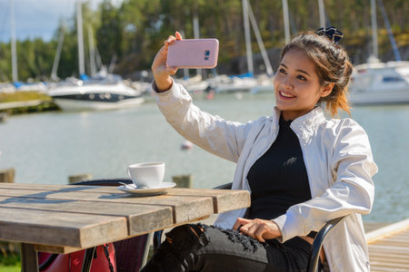 Happy young beautiful Asian tourist woman taking selfie at restaurant by the pierの写真素材