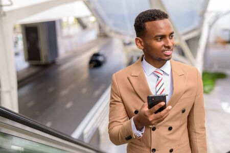 Happy young handsome African businessman using phone while moving up the escalator in the cityの写真素材