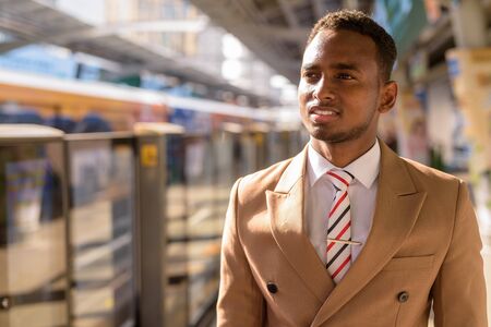 Happy young handsome African businessman thinking and waiting at the sky train stationの写真素材