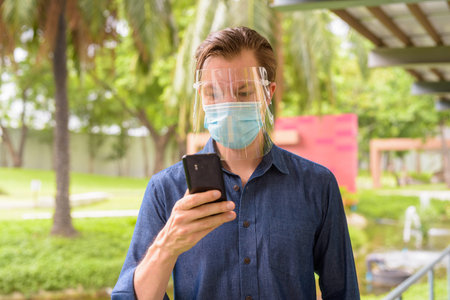 Face of young man using phone with mask and face shield for protection from corona virus outbreak at...