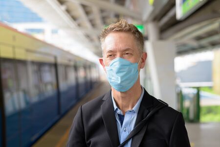 Face of mature businessman with mask thinking and waiting at the sky train stationの写真素材