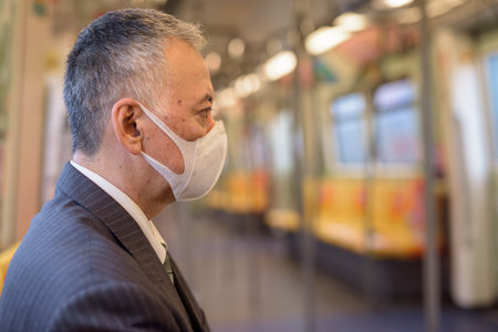 Mature Japanese businessman wearing mask and sitting with distance inside the train