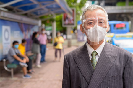 Mature Japanese businessman with mask and face shield at the bus stop